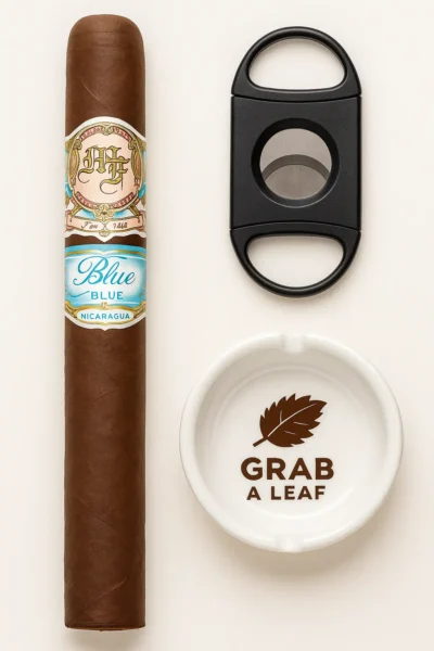 Full view of a premium My Father Blue cigar displayed vertically from head to foot, with a silver cigar cutter and branded ashtray beside it on a clean background.